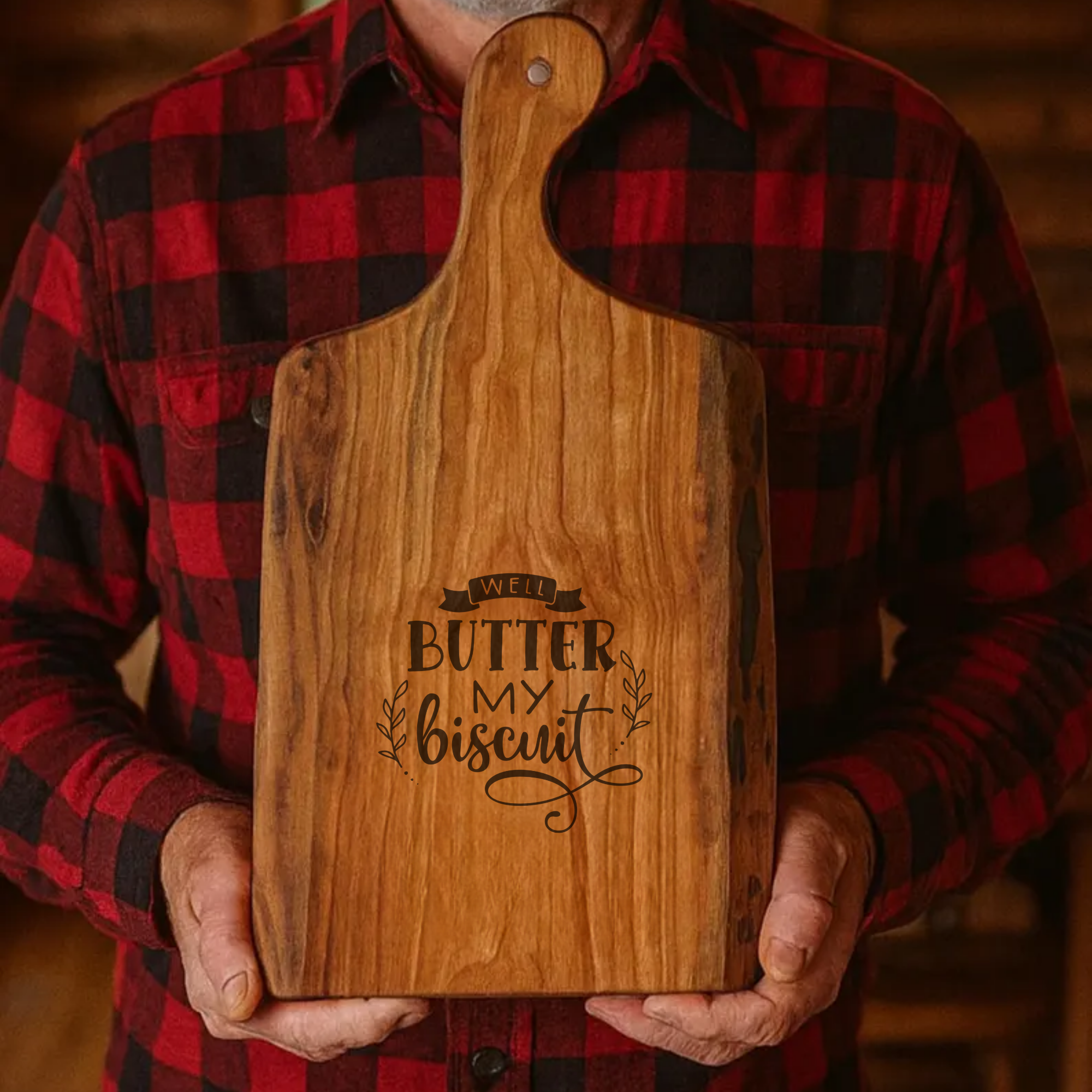 Person holding a wooden cutting board with 'Butter My Biscuit' engraving, wearing a red and black plaid shirt.