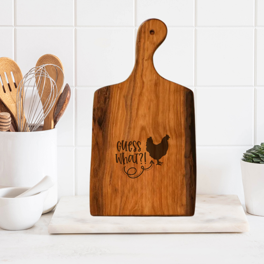 Wooden cutting board with 'Guess What?!' and a chicken illustration on a kitchen counter.