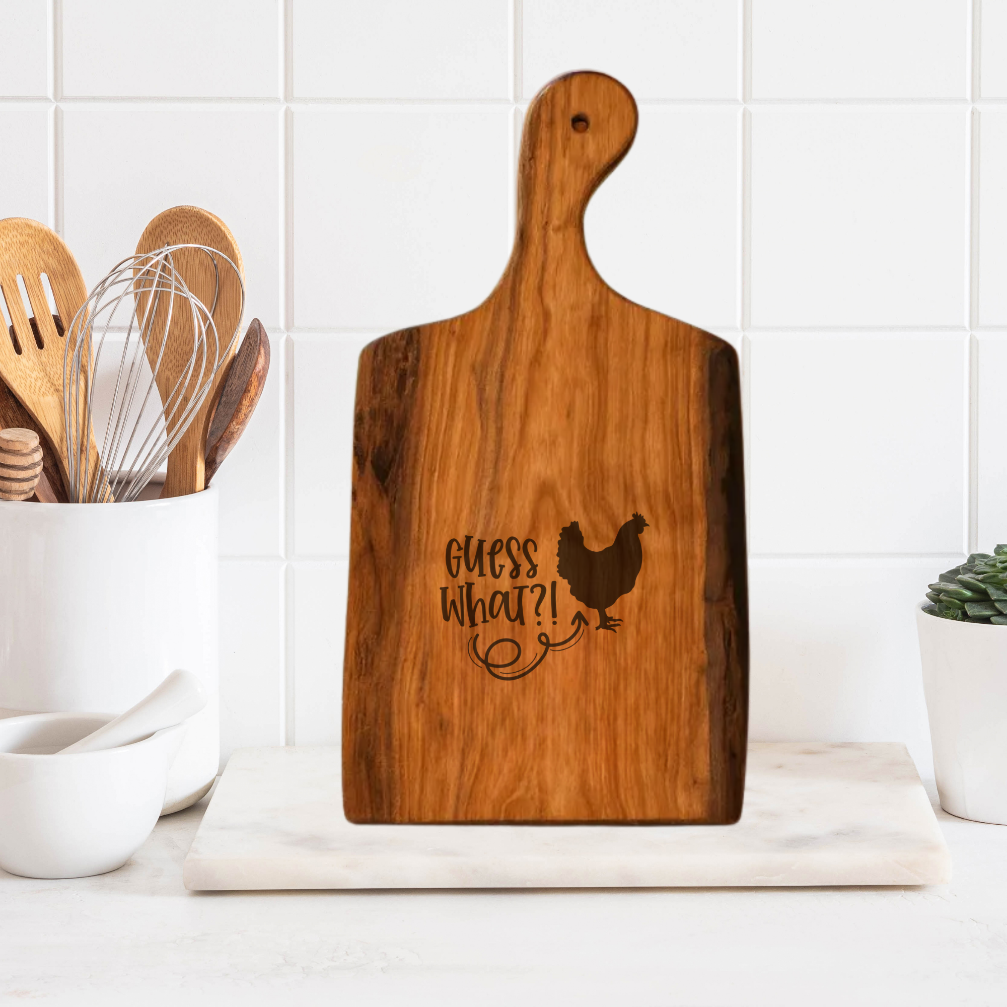 Wooden cutting board with 'Guess What?!' and a chicken illustration on a kitchen counter.