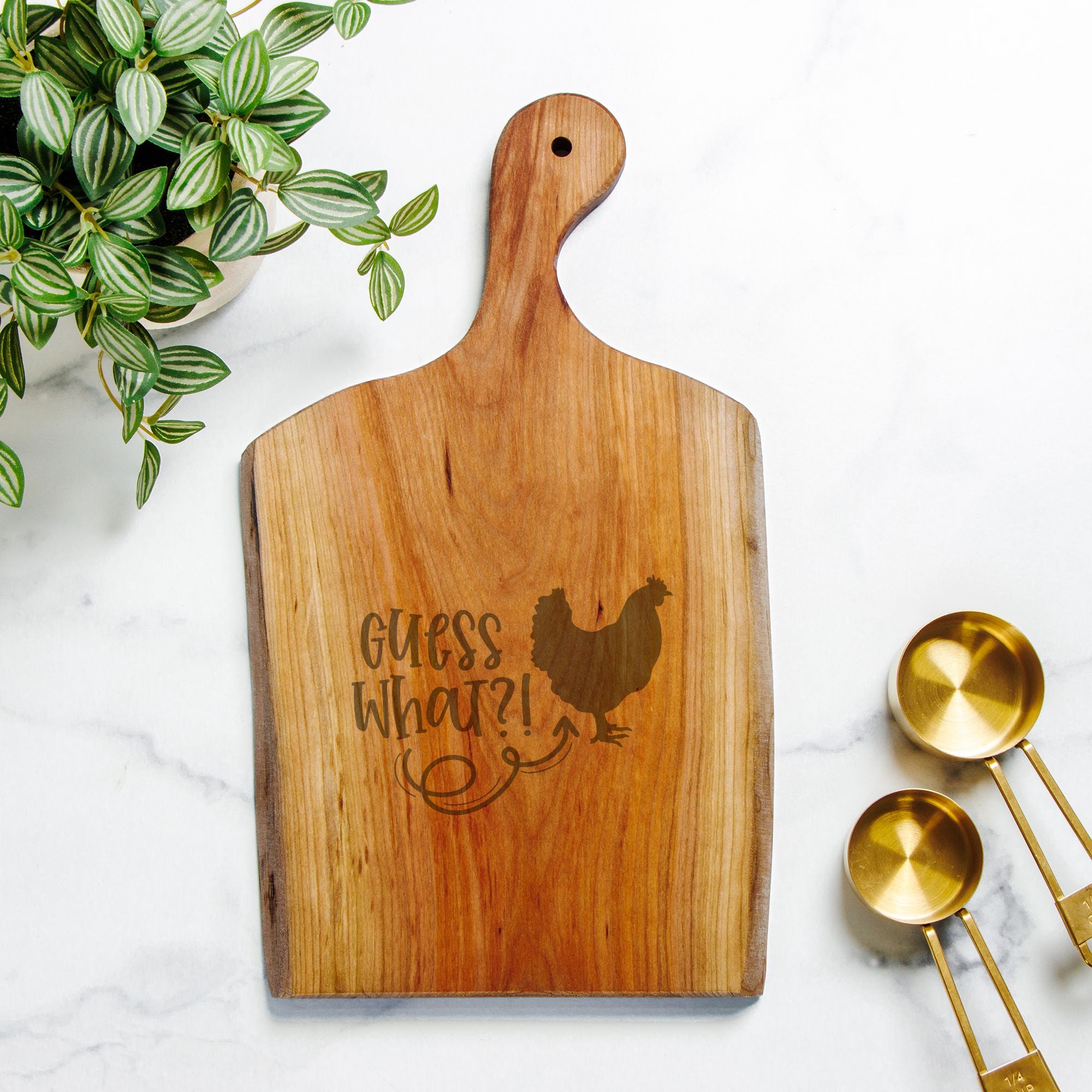 Wooden cutting board with engraved text and rooster design, placed on a marble surface with gold measuring spoons and a plant.