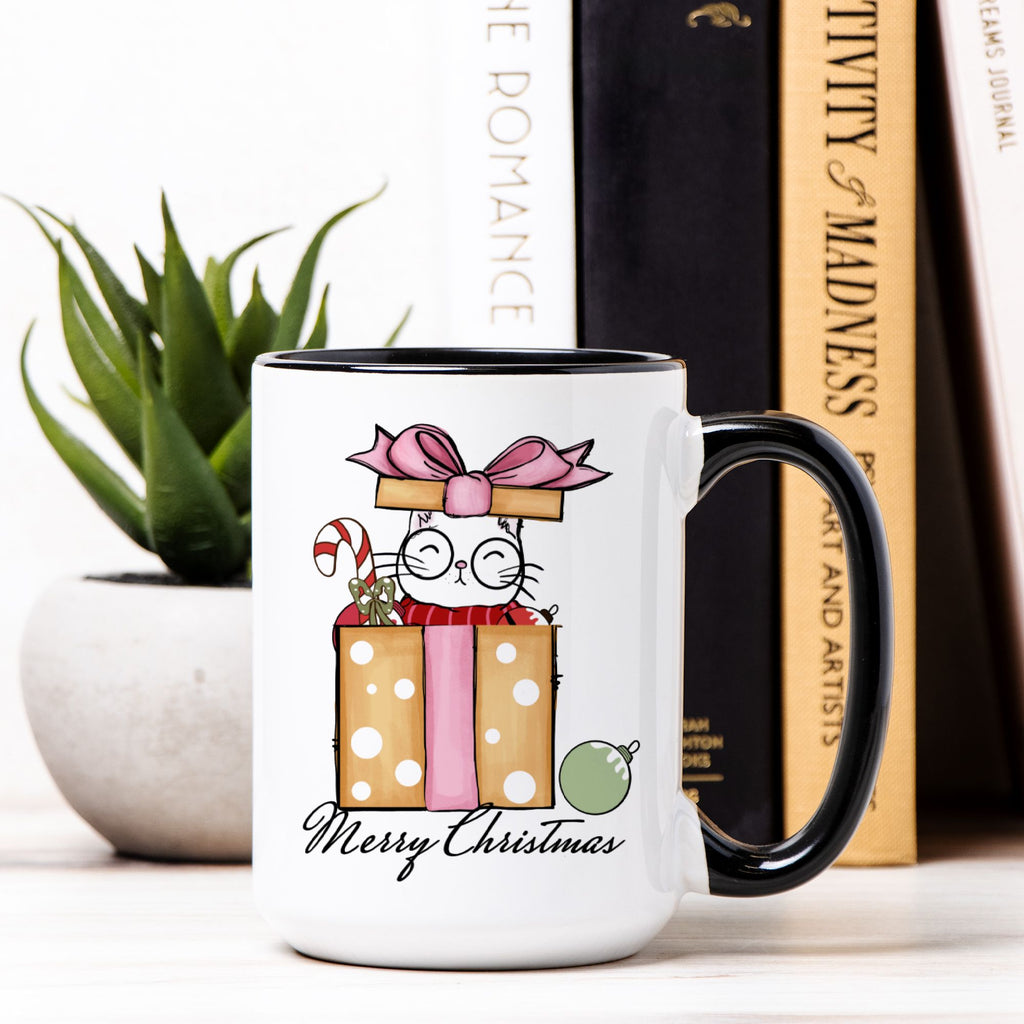 White mug with black interior and handle, featuring a Christmas-themed design of a cat in a gift box, on a surface with books and a plant.