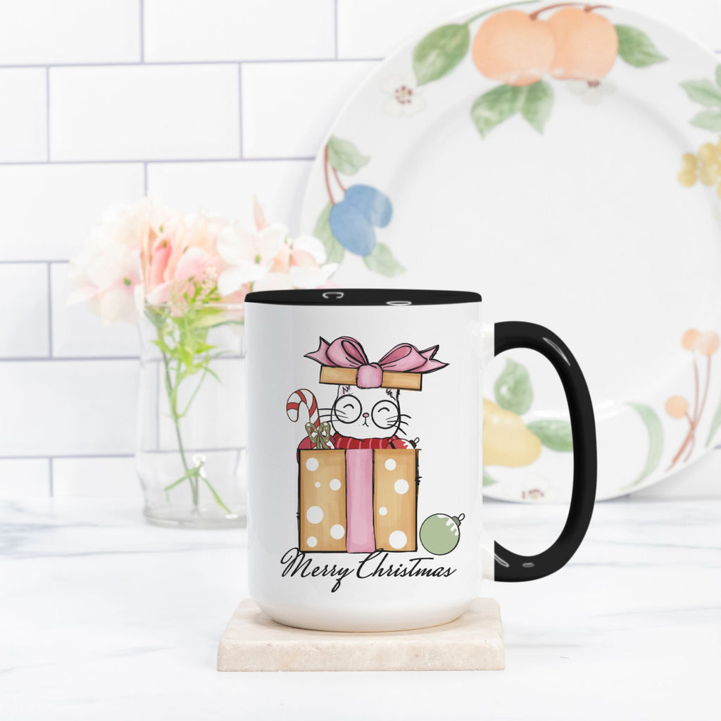 White mug with black handle featuring a Christmas-themed design of a cat with presents and 'Merry Christmas' text, placed on a white surface with a blurred background.