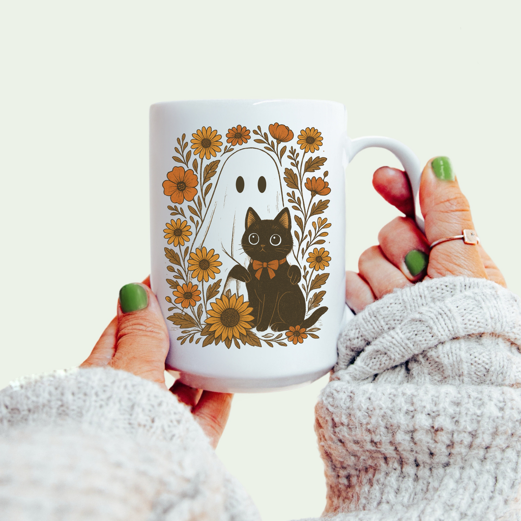 White mug with a black cat and floral design held by a person wearing a white sweater.