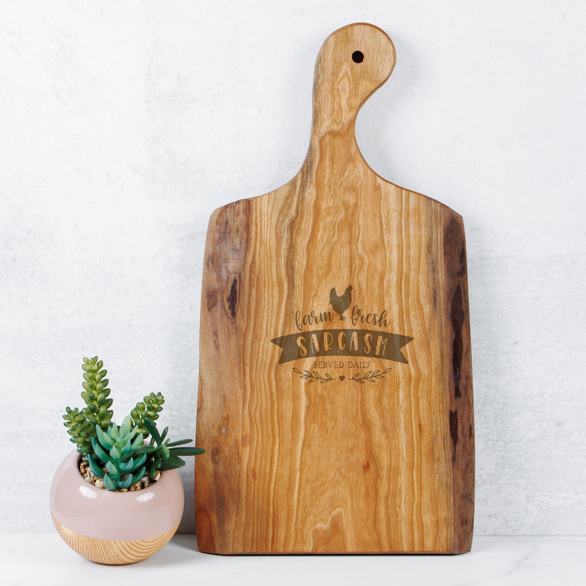 Wooden cutting board with engraved text next to a small potted plant on a light background