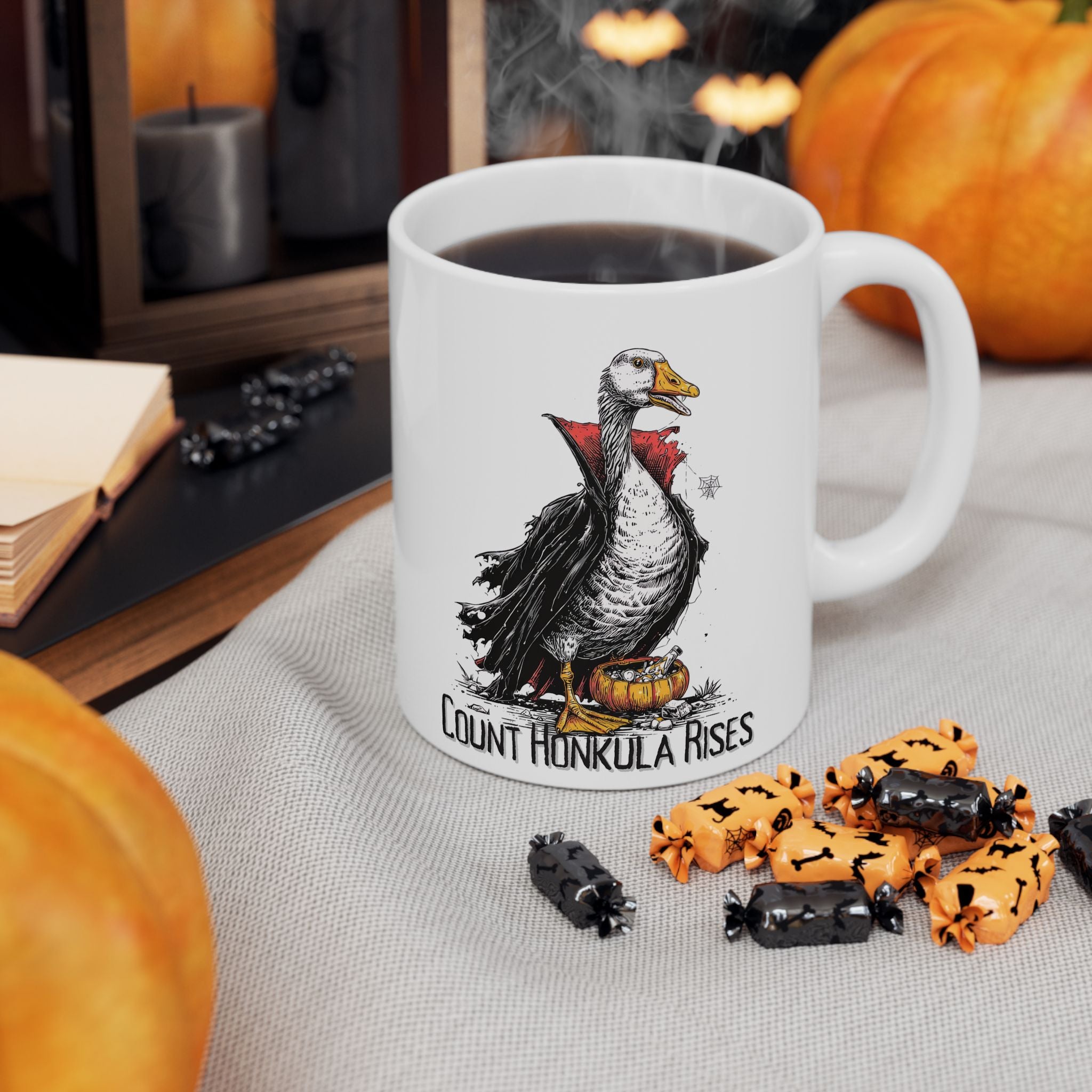 White mug with a vampire goose illustration and text 'Count Honkula Rises', surrounded by Halloween-themed treats and pumpkins.