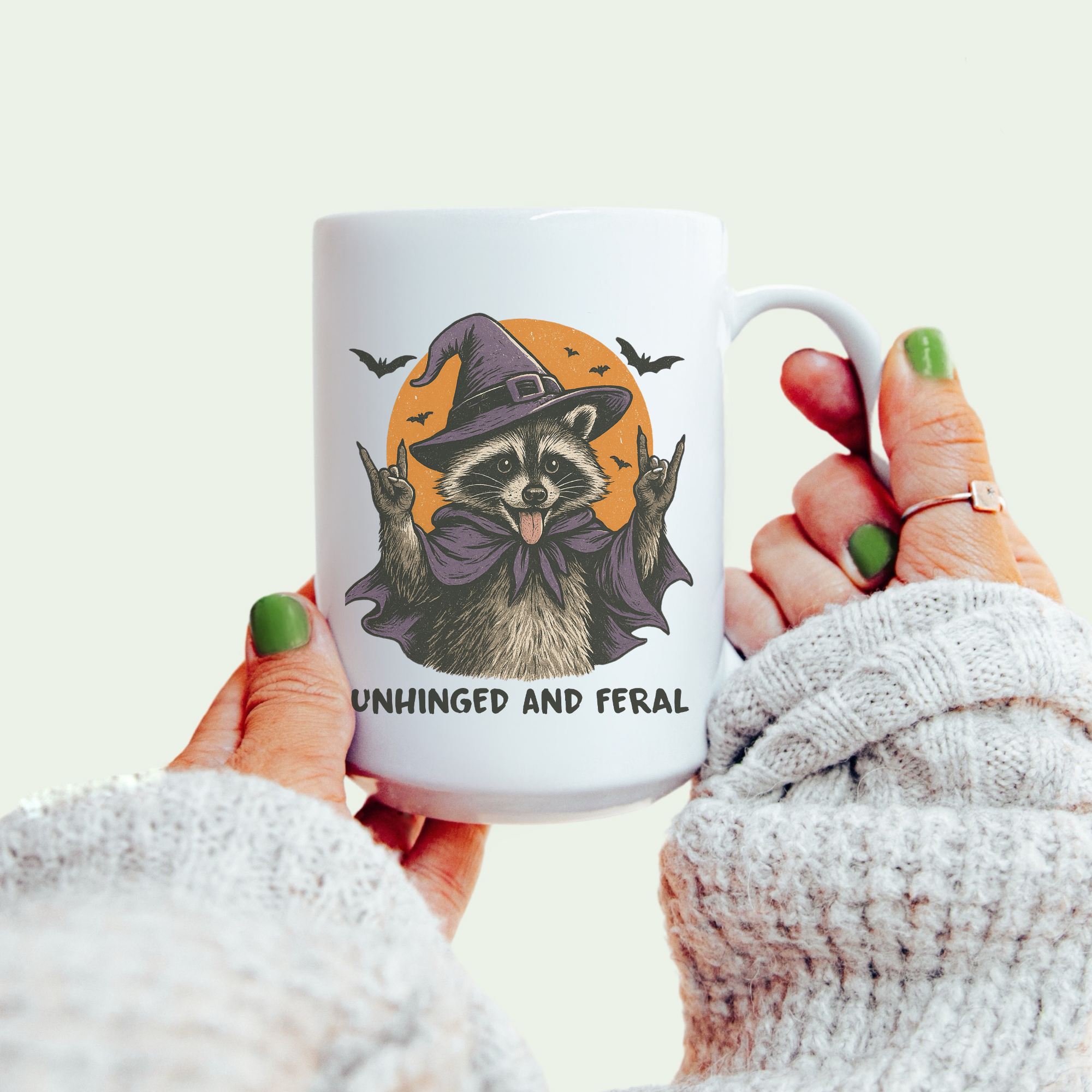 Mug with a raccoon wearing a witch hat design, held by a person with green nail polish.