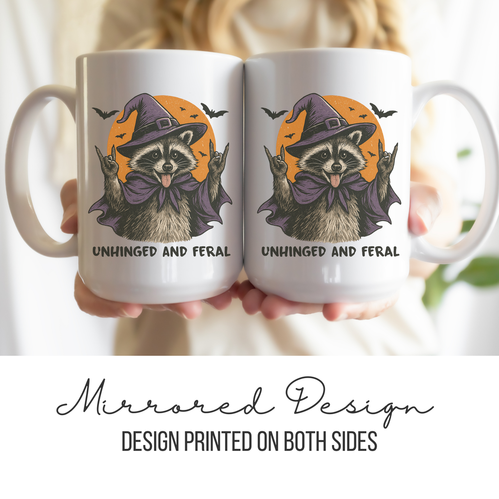 Two white mugs with a mirrored design of a raccoon wearing a witch hat and text 'Unhinged and Feral' held by a person.