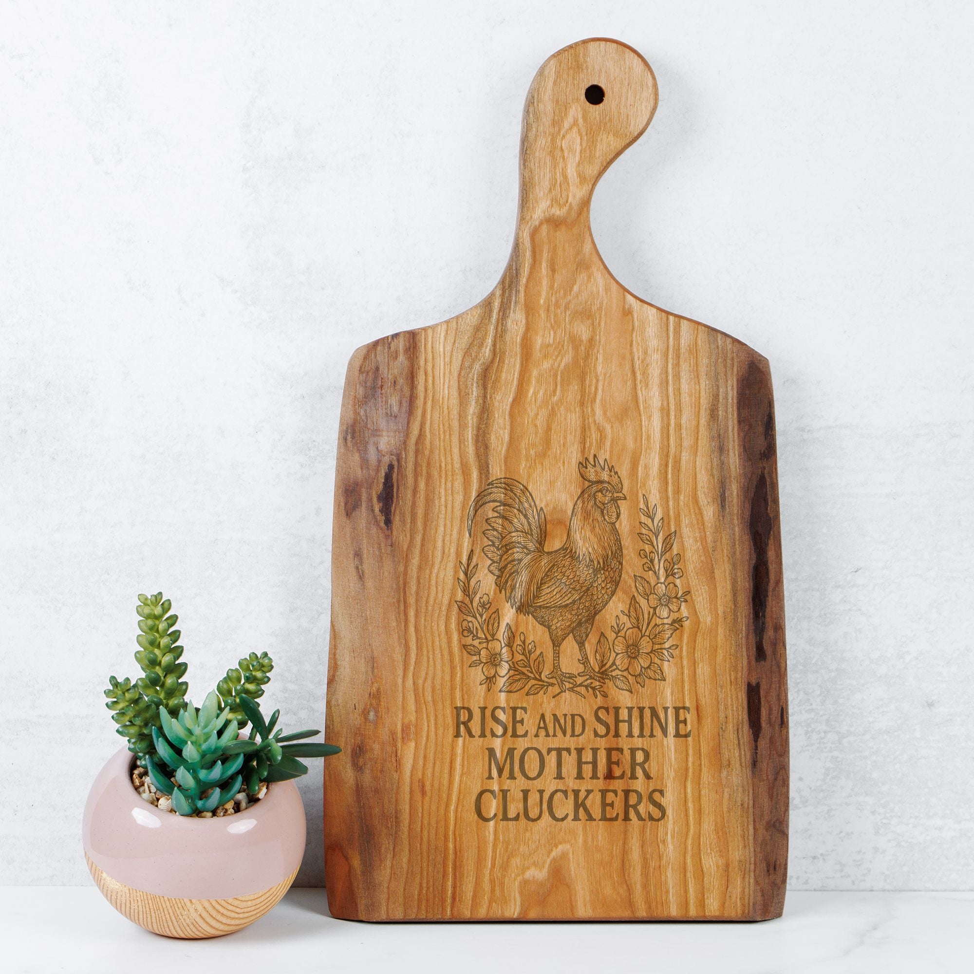 Wooden cutting board with rooster design and text next to a small potted plant on a light background