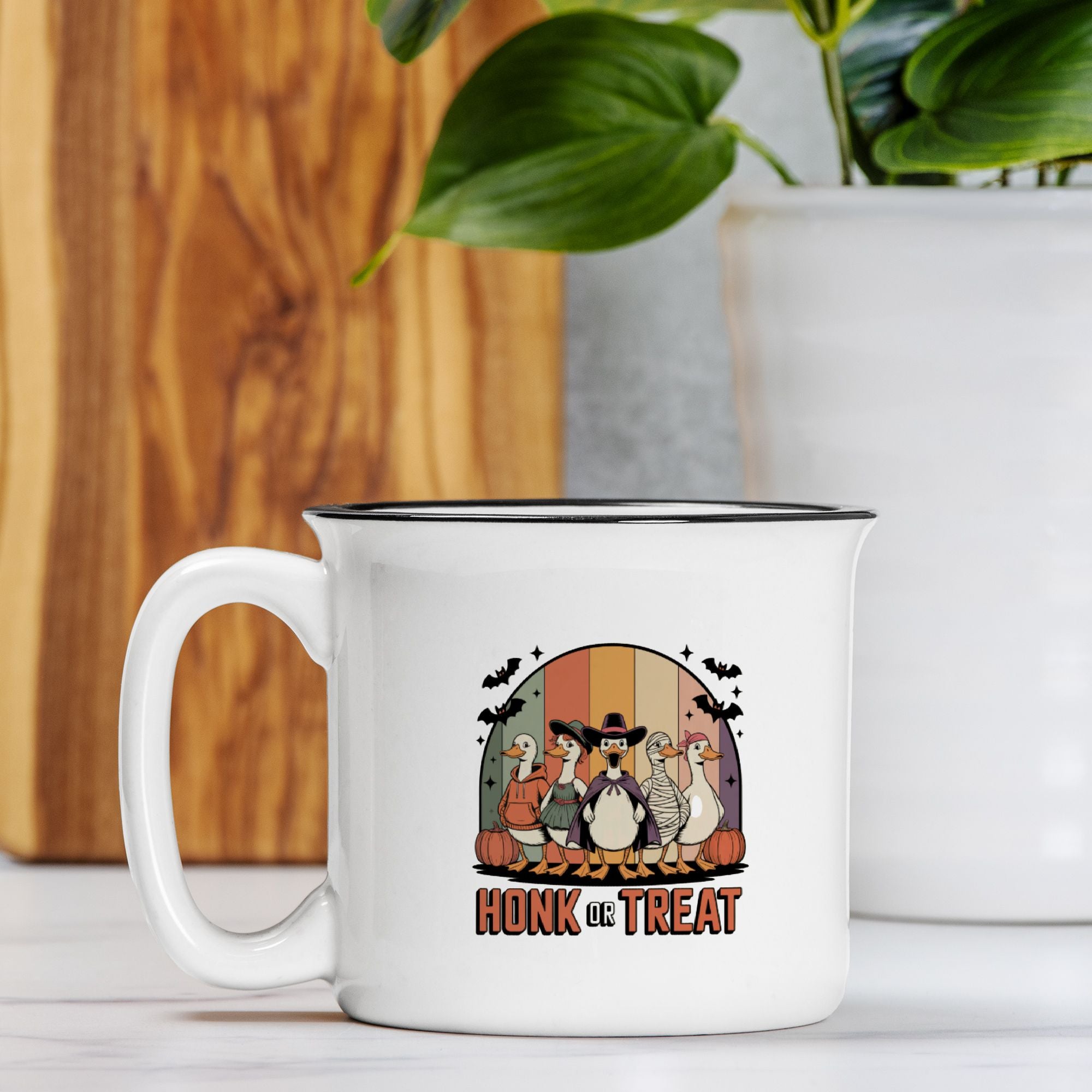 White camp mug with a Halloween-themed design featuring geese and text, placed on a wooden surface with a plant in the background.