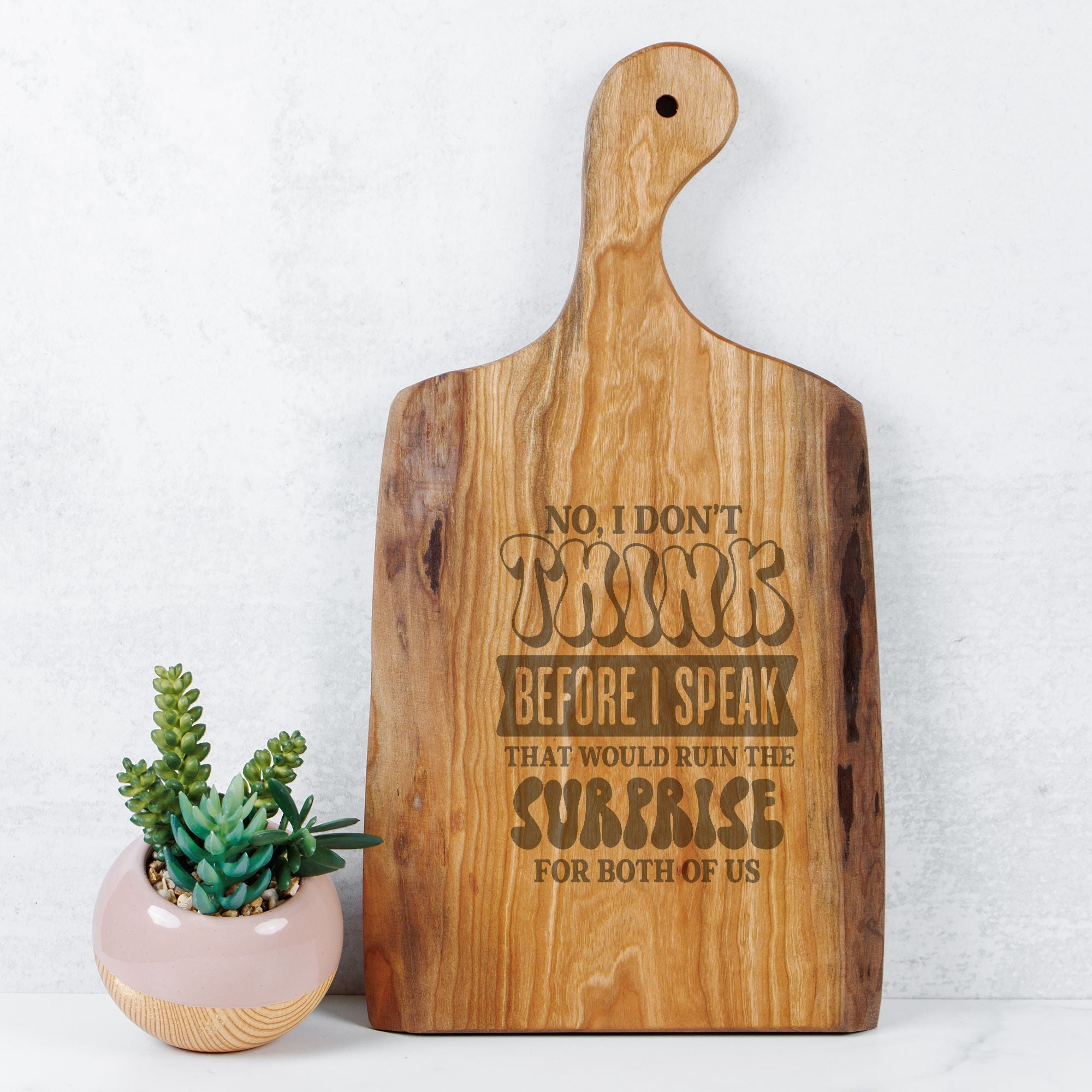 Wooden cutting board with engraved text next to a small potted plant on a light background