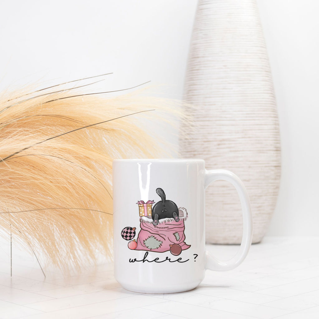 White mug with a cartoon design of a cat in a Christmas bag and 'where?' text, placed on a light surface with decorative elements.