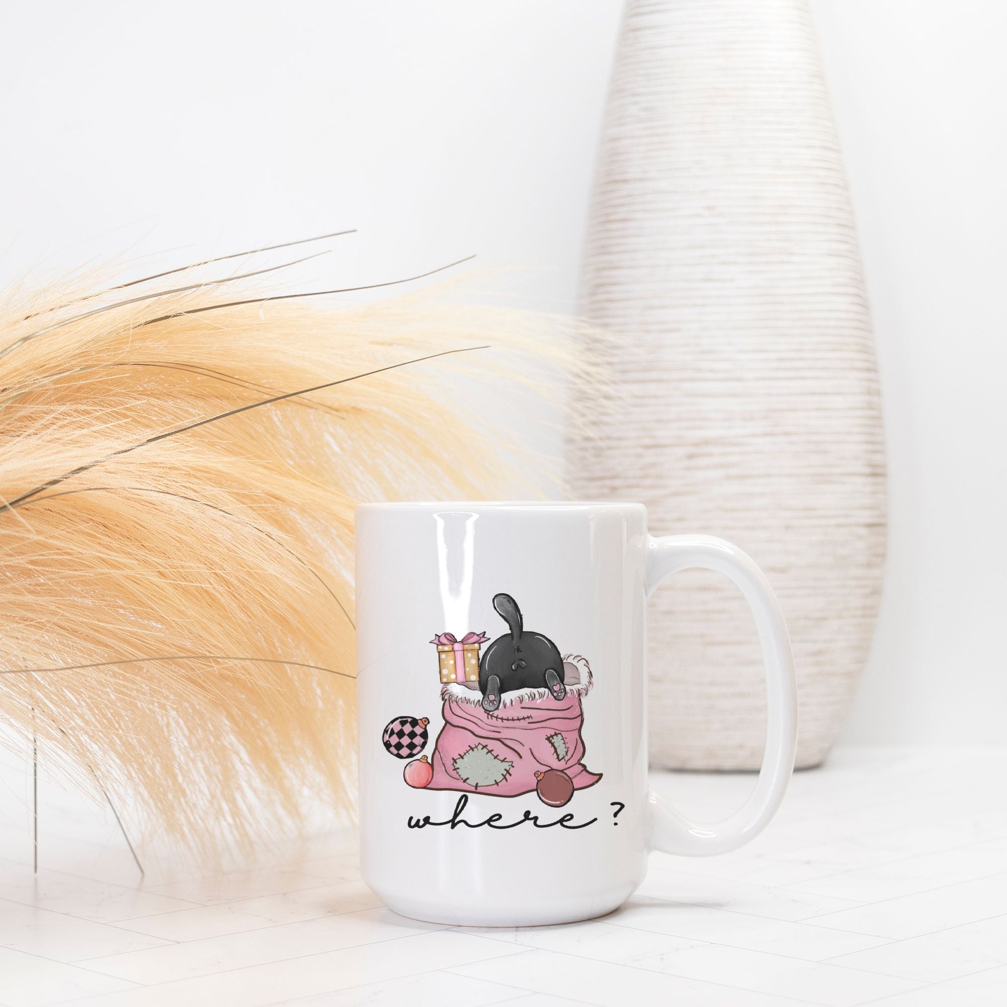 White mug with a cartoon design of a cat in a Christmas bag and 'where?' text, placed on a light surface with decorative elements.