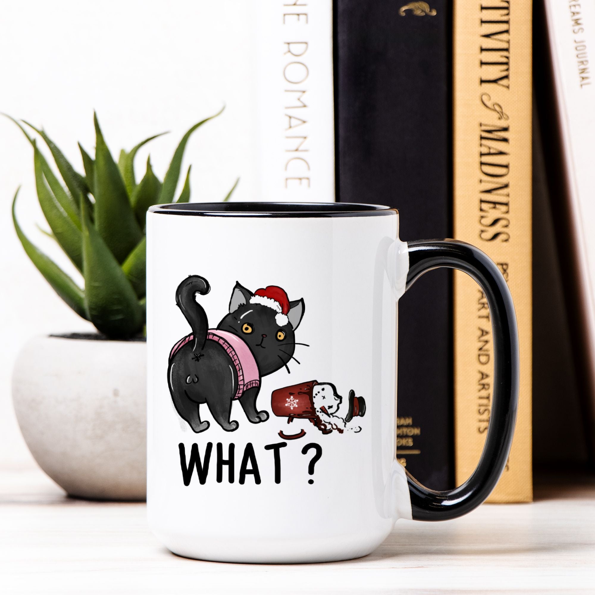 White mug with black interior and handle, featuring a cartoon Christmas cat, knocked over drink  and text 'What?', on a surface with books and a plant in the background.