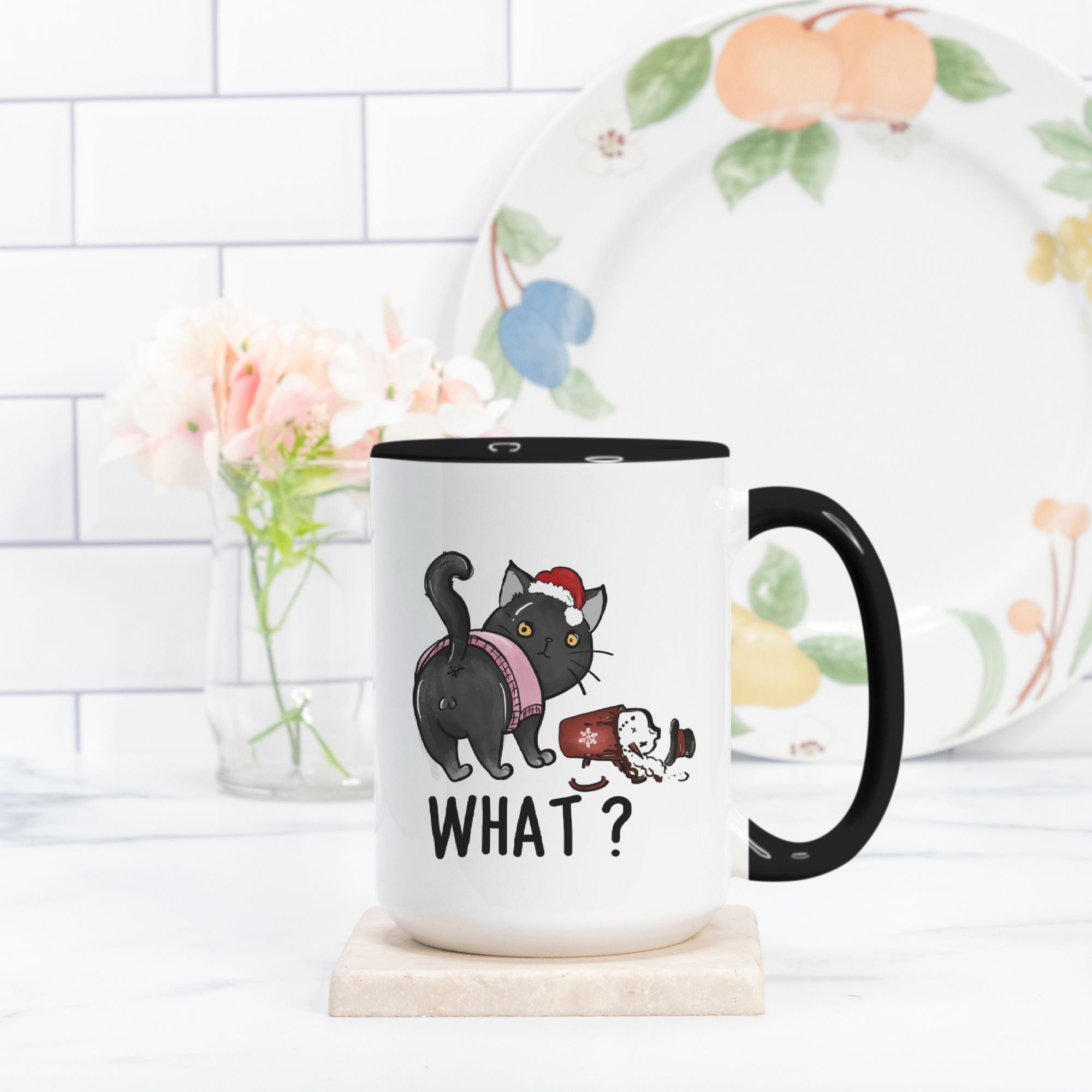 White mug with hand drawn black cat dressed for Christmas design and 'WHAT?' text, placed on a wooden coaster with a floral-patterned plate in the background.