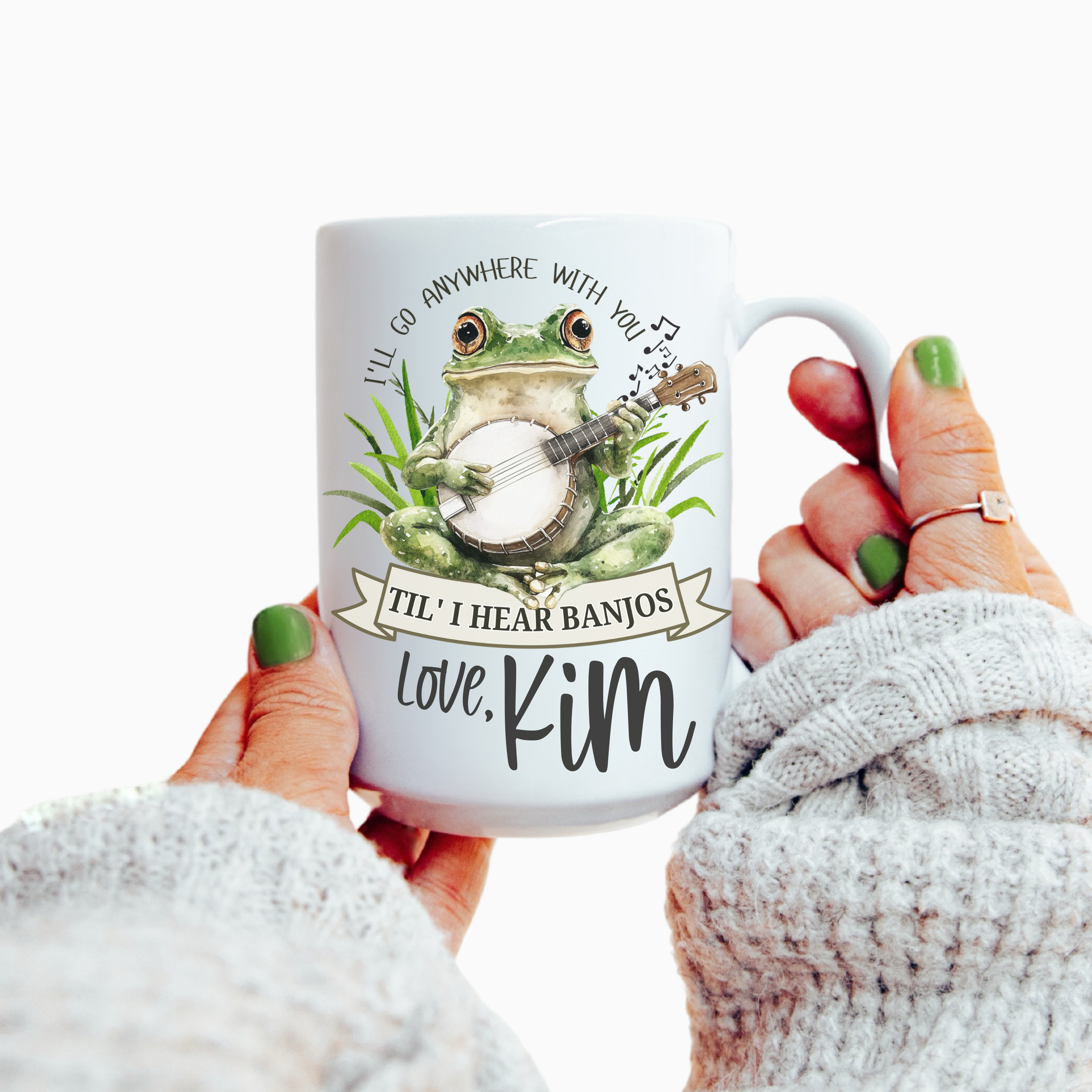 Mug with a frog playing a banjo design and text ' I'll go anywhere with you Til' I Hear Banjos', held by a person.