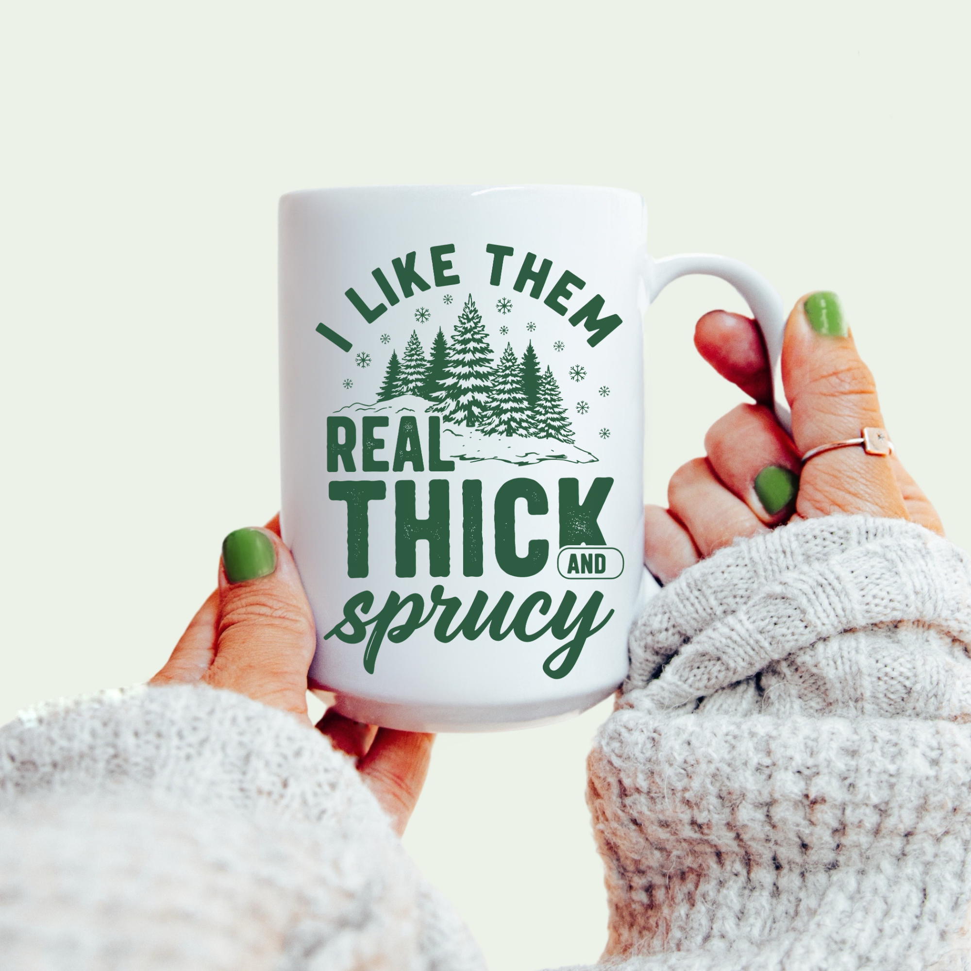 Mug with 'I Like Them Real Thick and Sprucy' text held by a person wearing a white sweater.