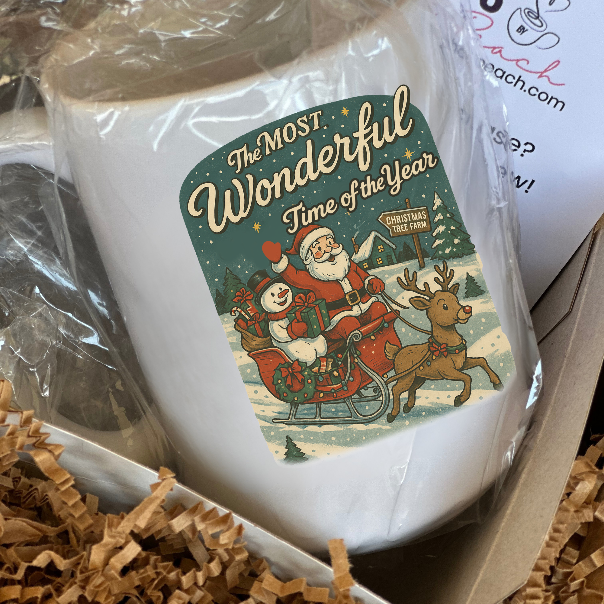 White mug with Christmas-themed design featuring Santa and reindeer, wrapped in clear plastic.