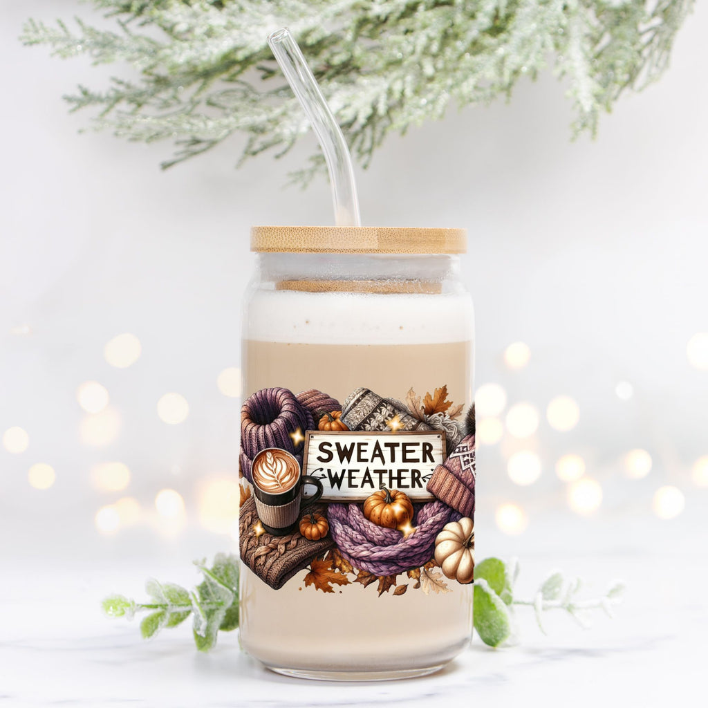 Glass jar with lid and straw featuring 'Sweater Weather' design on a white background with blurred lights and greenery.