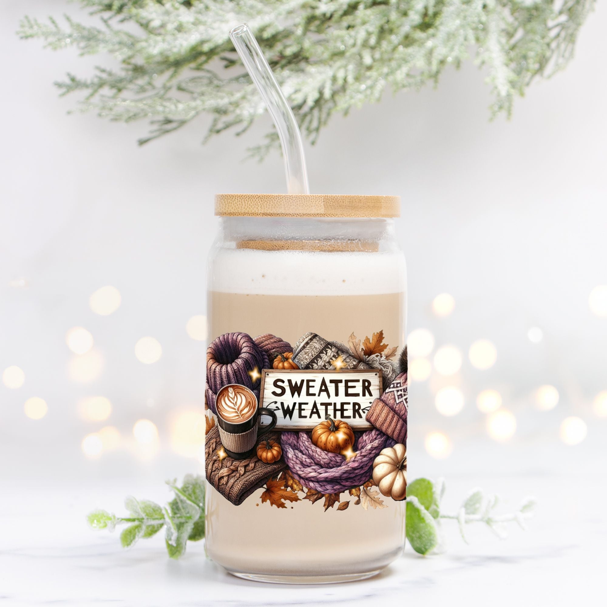 Glass jar with lid and straw featuring 'Sweater Weather' design on a white background with blurred lights and greenery.