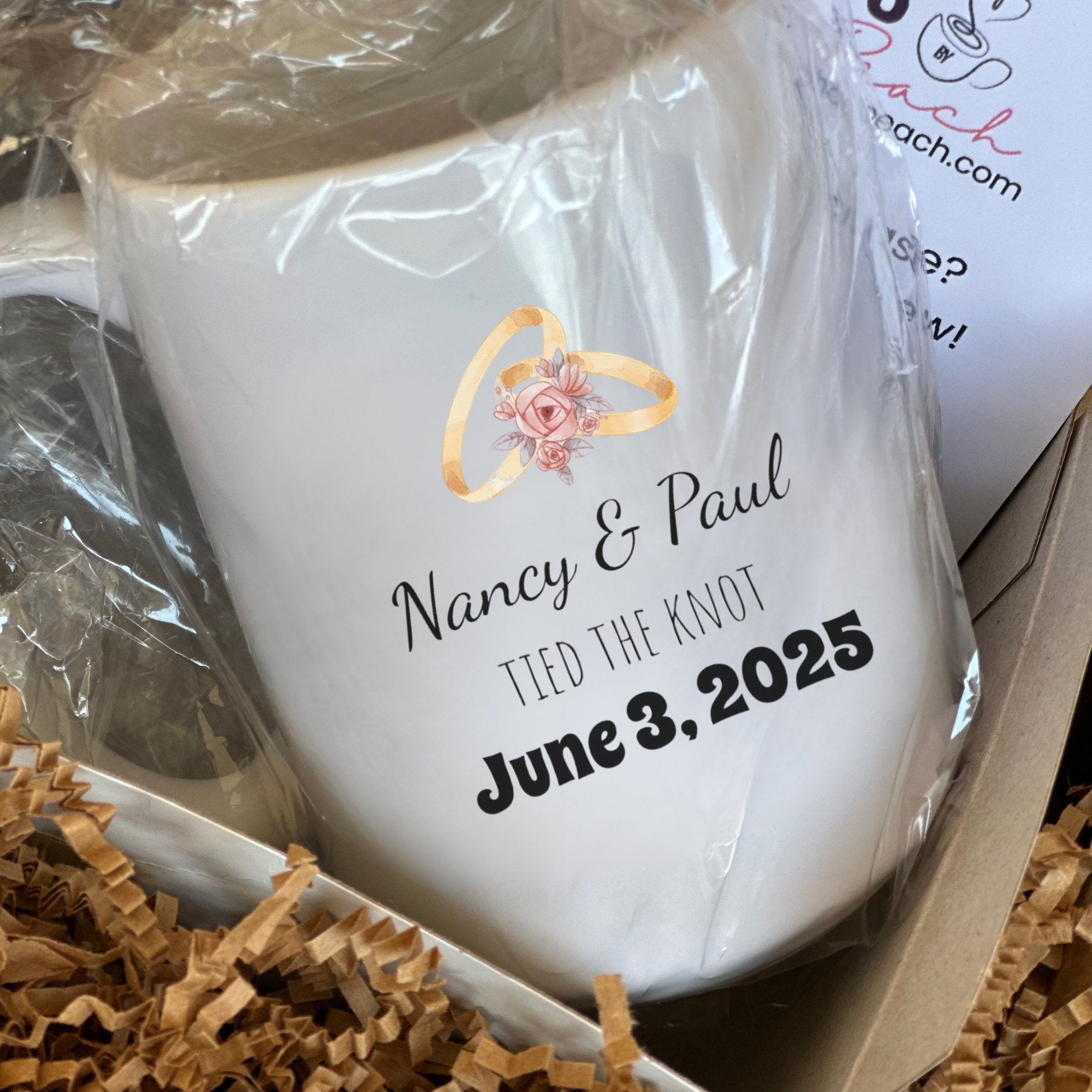 White mug with personalized text and ring design, wrapped in clear plastic, on a brown textured surface.