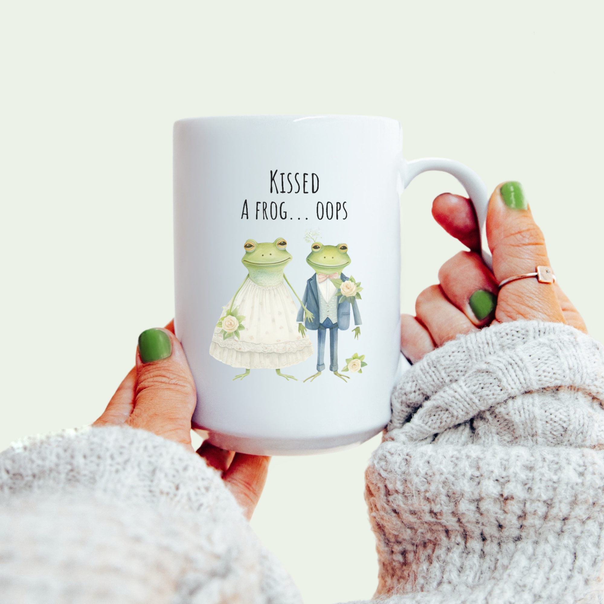 Mug with frog couple design and text held by a person wearing a white sweater.