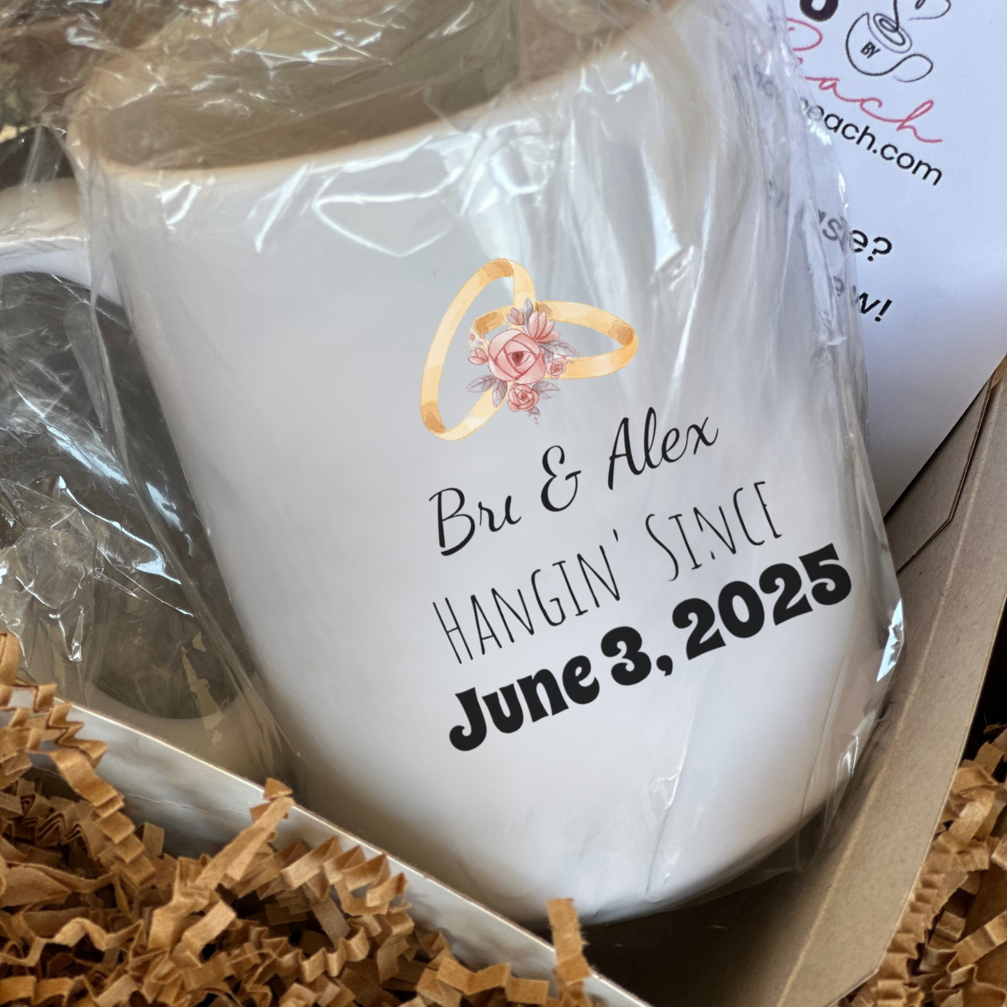 White mug with personalized text and ring design, wrapped in plastic, on a box with brown tissue paper.