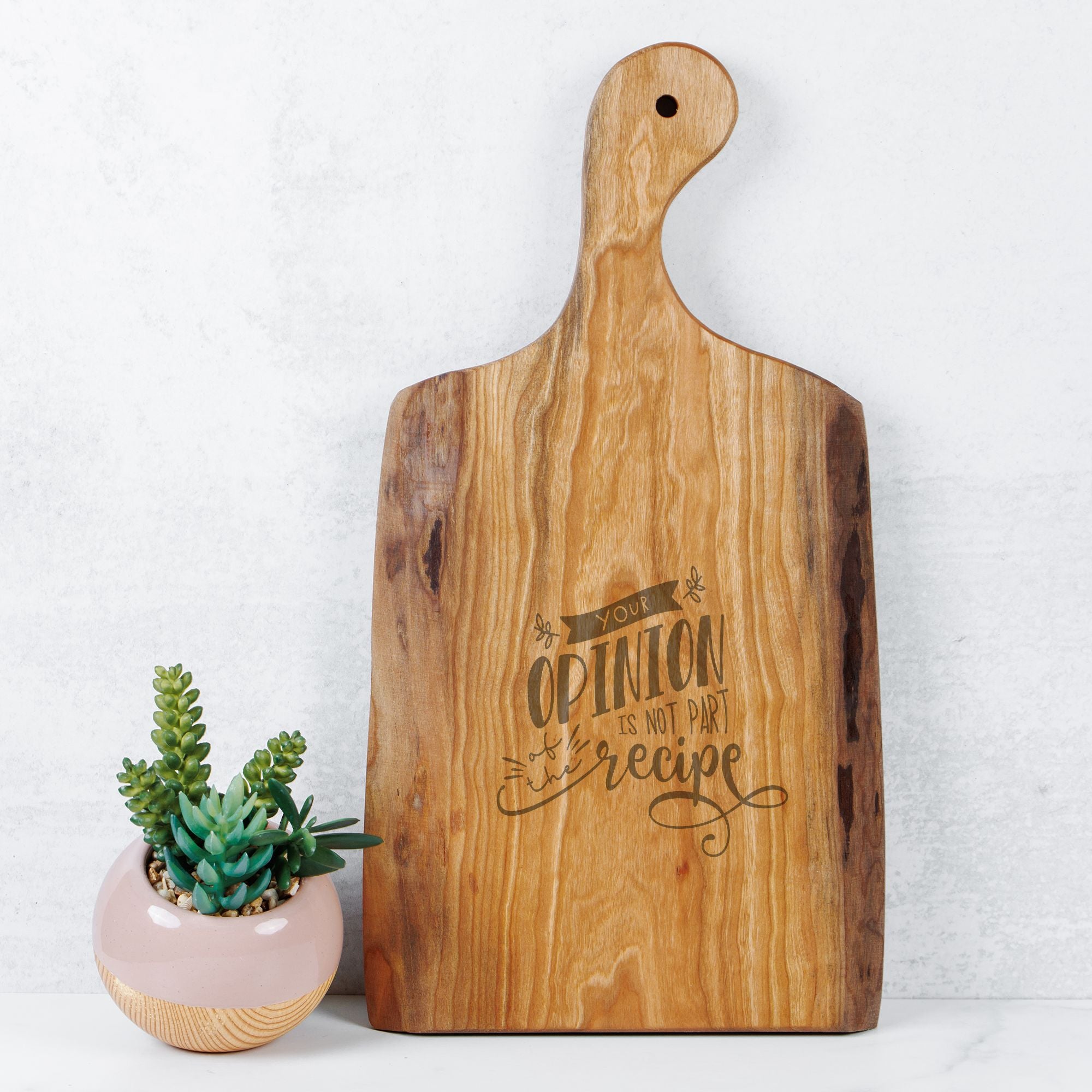 Wooden cutting board with engraved text next to a small potted plant on a light background
